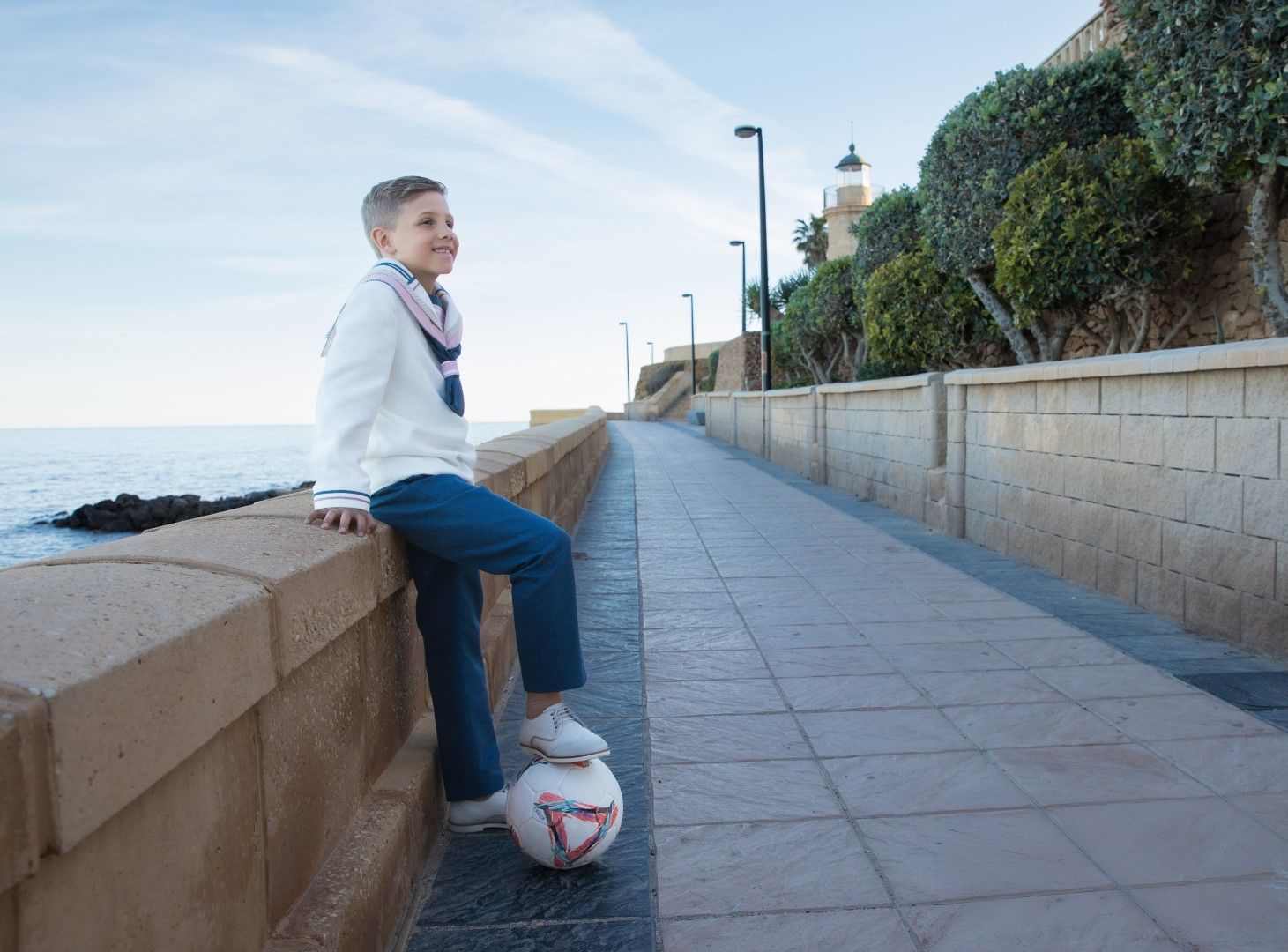 Niño de comunión sentado en el muro del paseo marítimo con un balón