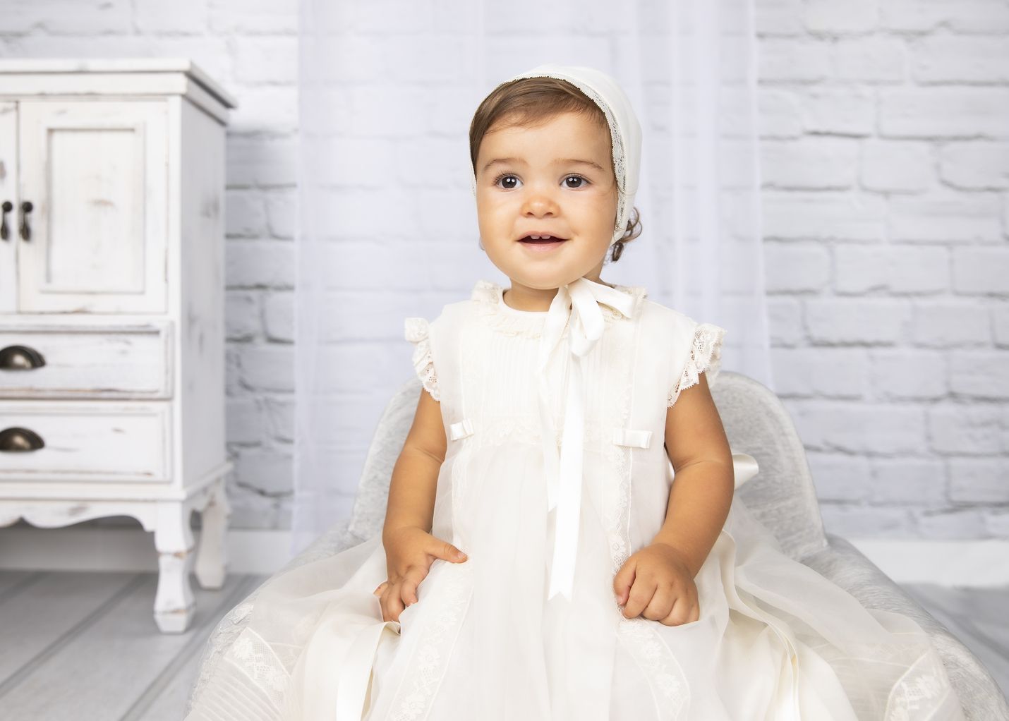 Bebé con vestido blanco junto a un mueble antiguo